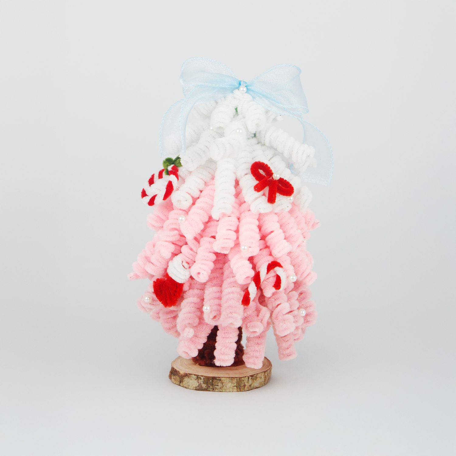Handmade Christmas tree made of pipe cleaners in pink and white with red accents, candy cane and bow decorations, blue ribbon on top, displayed on a wooden base.